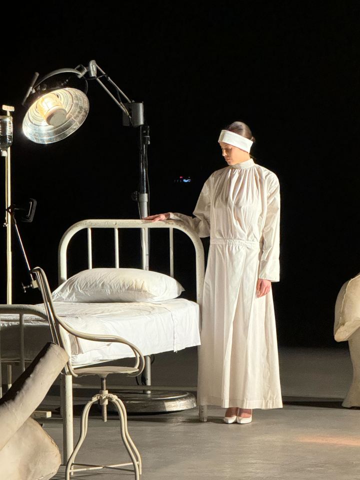 Woman in white robe standing by hospital bed.